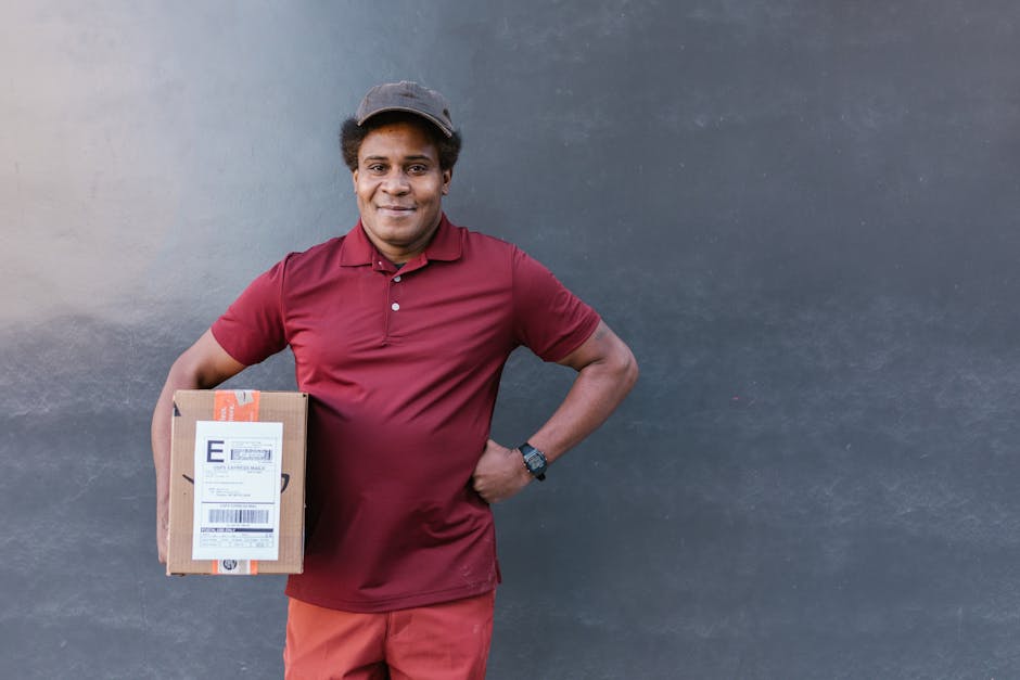 A smiling delivery person stands against a dark grey wall, holding a small cardboard package wrapped with orange and white labels. He is wearing a maroon polo shirt, matching maroon trousers, a grey c