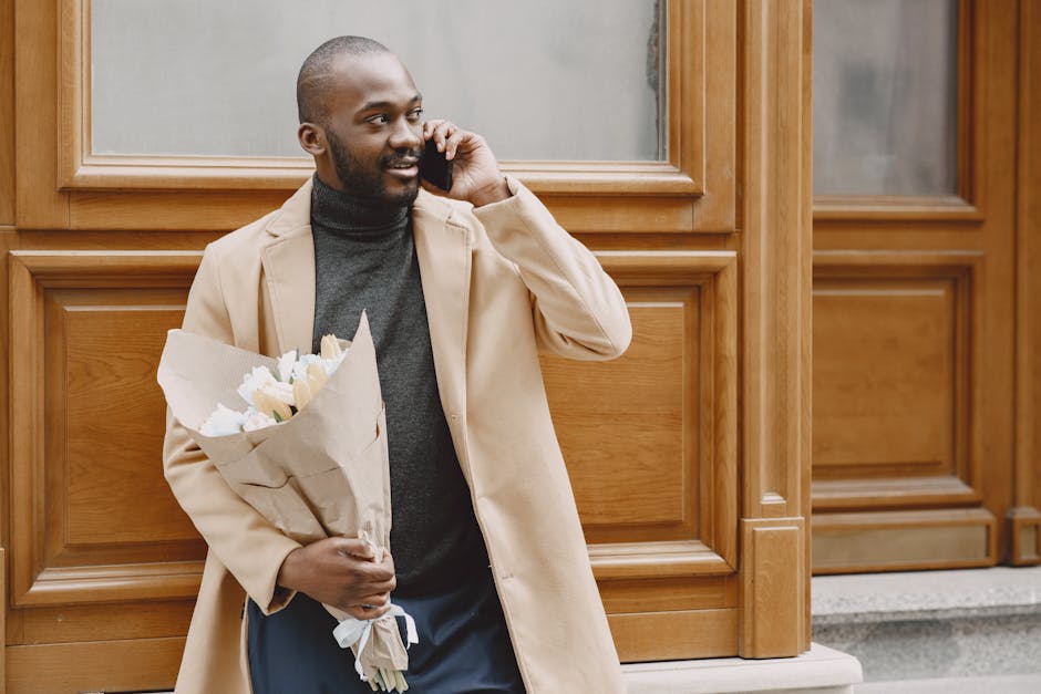 A man with a dark complexion and short hair, dressed in a beige coat and grey turtleneck, is standing outside against a wooden-paneled building while holding a bouquet of fresh white tulips wrapped in
