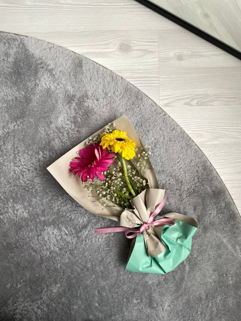 A vibrant floral gift featuring a small bouquet of fresh flowers, including two pink gerbera daisies, a bright yellow gerbera daisy, and delicate white baby's breath. The bouquet is wrapped in beige a