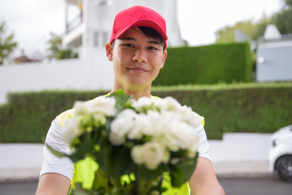 A smiling young male flower delivery courier wearing a red cap and high-visibility vest is standing outdoors on a residential street, holding out a bouquet of fresh white roses and calla lilies wrappe