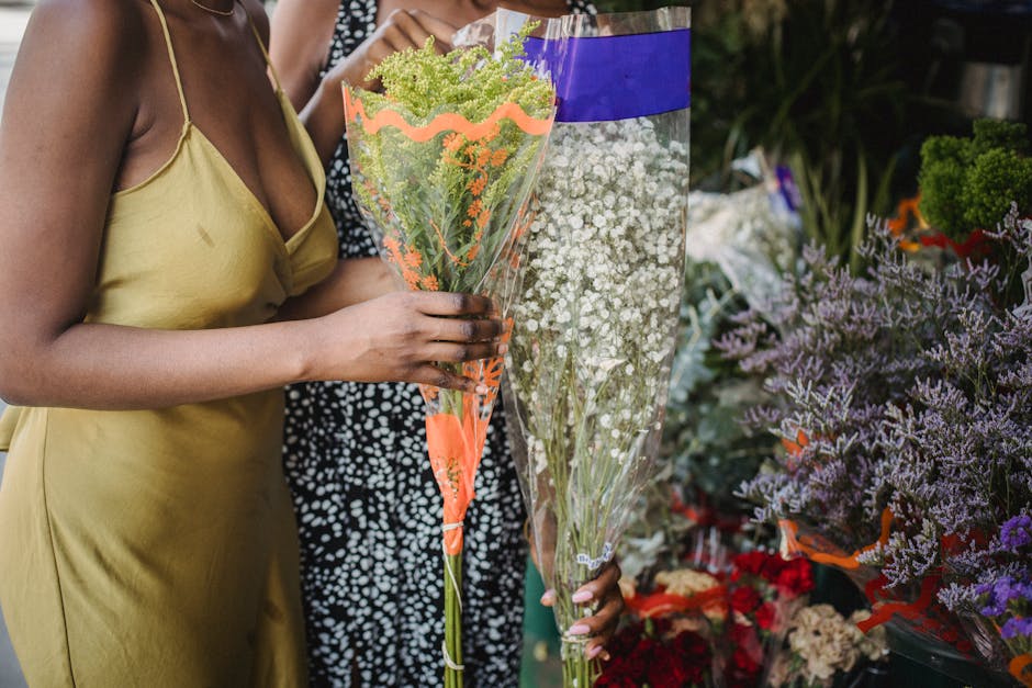 A person wearing a yellow dress holds a large bouquet of fresh, vibrant green and purple flowers wrapped in clear plastic with orange and purple accents, standing next to another bouquet of small whit