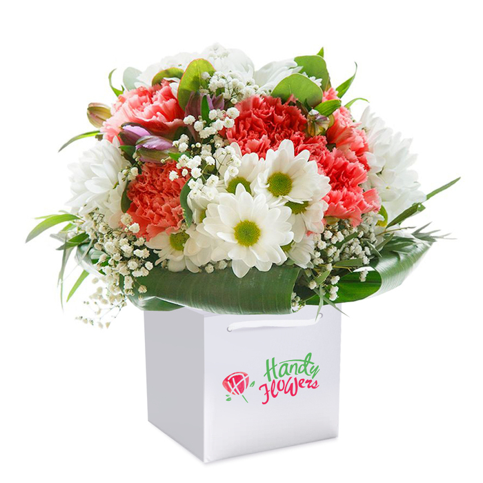Displayed in a pristine, square white gift bag set before a flawless white background, this bouquet exudes classic charm with a modern twist, the sort often found in thoughtful gifts exchanged between Harold Wood neighbours. The main event is a lush cascade of ruffled carnations in the softest coral and peach, punctuated by vivid reds that recall sunsets over Pages Wood. Dancing throughout the design, pure white daisy-like chrysanthemums with their fresh lime-green hearts add a note of playful serenity. Delicate baby's breath weaves between the main blooms, its tiny white flowers creating clouds of gentle texture and amplifying the sense of airiness. Just off to one side, a handful of unopened buds tinged purple invite closer inspection, deepening the arrangement with their quiet promise. At the base, a collar of bold, dark green foliage encircles the flowers-each leaf glossy and substantial, reflecting the florists' careful hand. The sturdy ribbon handles on the bag convey its suitability for easy gifting and delivery, perfect for birthdays, anniversaries, or spontaneous gestures of kindness around Harold Wood. The careful composition and evident freshness make this arrangement a natural fit for the area's discerning flower lovers, with every detail capturing not just beauty but also the spirit of thoughtful local connection.