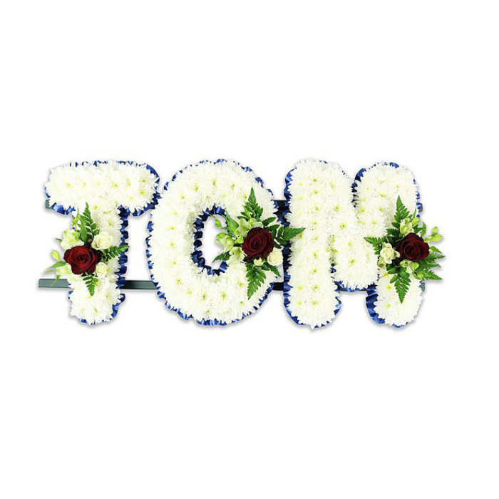 A dignified, personalised floral name tribute that spells TOM in large, sculptural letters, each formed from a dense carpet of small, snow-white chrysanthemums whose layered petals create a plush, tactile surface. The arrangement is crafted to read clearly from a distance: every chrysanthemum is placed to preserve an even, rounded profile while allowing soft shadow and depth to show beneath the petals. The outline of each letter is finished with a vivid royal blue ribbon that crisply defines the silhouette and lends an element of ceremonial formality, the blue catching light differently from the matte white blooms. Placed deliberately within the centre-to-lower portion of each letter is a compact floral spray dominated by a single deep red rose-its velvet texture and subtle perfume symbolise affection and remembrance-surrounded by smaller creamy-white spray roses and neat buds which ease the transition from intense red to calm white. Fresh fern fronds and a mixed selection of glossy foliage provide verdant structure and contrast, their fine fronds and broader leaves creating pockets of shadow and natural movement. A thin dark green or black metal support bar is visible behind the base, providing stability and hinting at professional workshop construction while keeping the flowers visually elevated against the crisp white backdrop. Prepared with the sensitivity of a local florist, this piece is suitable for funeral viewings, memorial gatherings, or graveside services around Harold Wood and nearby Romford; Flower Delivery Harold Wood can recreate this bespoke tribute with the same careful placement and respectful tone for families requesting local delivery or hand-assembled memorial pieces.