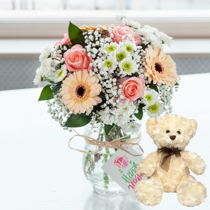 On a pristine white surface that catches and reflects the soft daylight, a charming anniversary gift pairing is carefully arranged, evoking the cosy, lived-in feel of Harold Wood homes. At the centre, a clear glass jar-style vase stands modestly, filled with water and visible stems. From this simple base blooms a generous, rounded bouquet in gentle pastel tones. Blush pink roses with velvety petals nestled close, warm peach gerbera daisies with distinctive dark brown centres, and clusters of crisp white chrysanthemums with subtle lime-green centres combine to create a balanced, harmonious palette. Threaded throughout are fine, cloud-like sprays of white baby's breath, which soften the outlines of the bouquet and give it an almost romantic haze. A few sprigs of dark green foliage peek through, ensuring the arrangement feels fresh and garden-inspired. Around the vase neck, a strand of natural twine is tied in an understated bow, from which hangs a small rectangular white tag. This tag displays a simple pink heart design at the top, with the word Handmade rendered in graceful green script, suggesting the thoughtful touch of a local Harold Wood florist familiar with marking milestones and anniversaries in the community. To the right of the vase sits an inviting cream-coloured teddy bear, upright and slightly turned toward the flowers. Its fur is soft and plush, with dark brown eyes and nose that give it a gentle, reassuring expression, and a neat brown satin ribbon bow around its neck that adds a classic, gift-worthy finish. The background is softly out of focus, filled with bright, diffused light, letting the flowers and bear remain the clear focal point. The whole scene feels intimate and affectionate, perfect for celebrating an anniversary, welcoming a new baby, or sending a comforting gesture across Harold Wood.