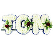 The photograph shows a traditional funeral flower tribute spelling out the name TOM in large capital letters, positioned side by side on a slender dark frame as though placed at the front of a service in Harold Wood. Each letter is thoroughly carpeted with white chrysanthemums, the blooms pressed closely together so their small, layered petals form a smooth, plush surface that covers every part of the backing. Running along the edge of each letter is a royal blue satin ribbon, folded into subtle pleats so it sits slightly raised, giving the tribute a neat, tailored outline and setting the white flowers in sharp relief against the plain, bright background. At selected points on the letters, small yet striking floral sprays break through the sea of white: deep red roses with velvety petals, accompanied by petite white or cream spray roses and slim white blossoms similar to freesia. These are surrounded by a halo of greenery-fern fronds with delicate frills and other fresh leaves-that brings texture and movement to the structured design. The resulting palette of white, blue, and red carries a feeling of respect, love, and serene remembrance. This kind of personalised name tribute, easily imagined as being arranged by Flower Delivery Harold Wood for a local family, offers a clear visual focus for mourners. It allows the name Tom to be seen and honoured in flowers, a tender and dignified symbol placed close to the coffin or near the altar as friends and relatives gather to say farewell.